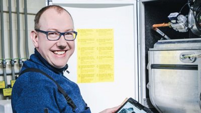 Ein lächelnder Techniker steht vor einer Maschine und hält ein Tablet in der Hand, während er auf eine gelbe Warnmeldung schaut.