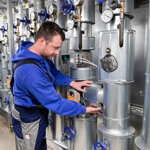 Ein Techniker in blauer Arbeitskleidung überprüft sorgfältig eine metallene Rohrleitung in einer industriellen Umgebung.