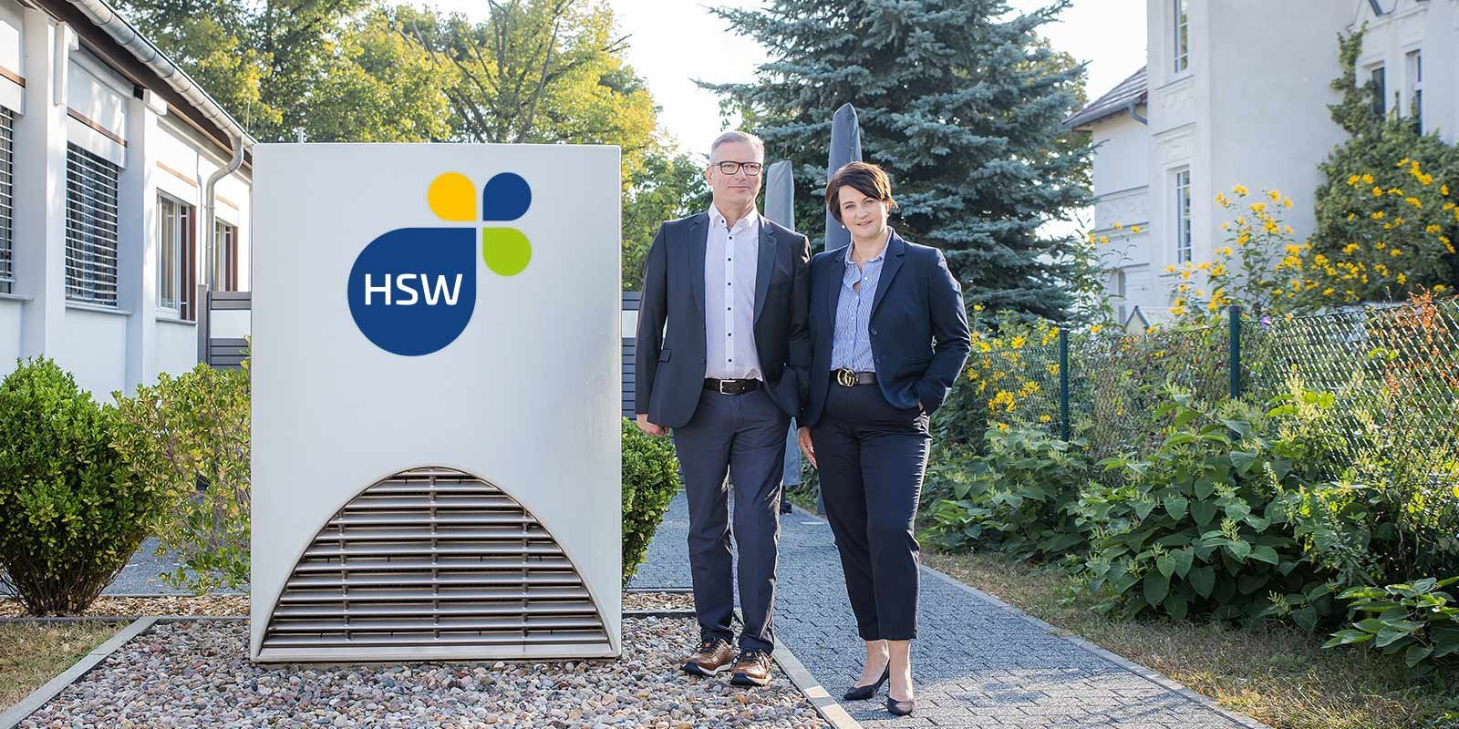 Ein Mann und eine Frau in formeller Kleidung stehen vor einem modernen Gebäude mit einem großen HSW-Logo.