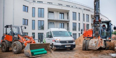 Auf der Baustelle stehen mehrere Baumaschinen, darunter ein Bagger und ein Lieferwagen, vor einem modernen Gebäude.