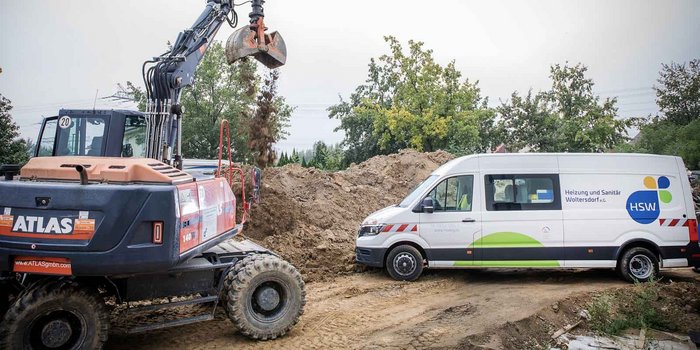 Ein Bagger hebt Erde an, während ein HSW-Fahrzeug in der Nähe parkt, um die Baustelle zu unterstützen.